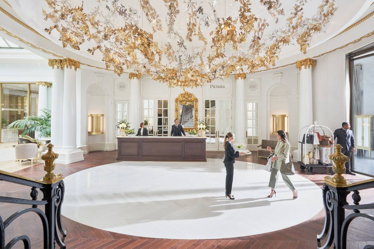 A grand lobby with a large white reception desk, ornate ceiling decorations, and classical architecture.