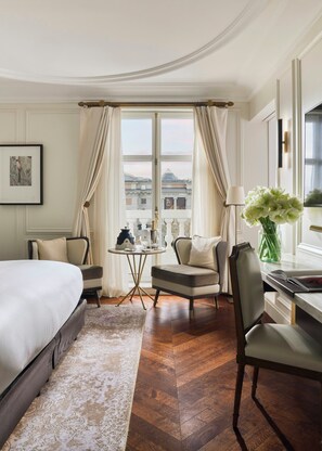 A hotel room with a bed, two armchairs, a desk, a lamp, a vase with flowers, and a view of buildings through large windows.