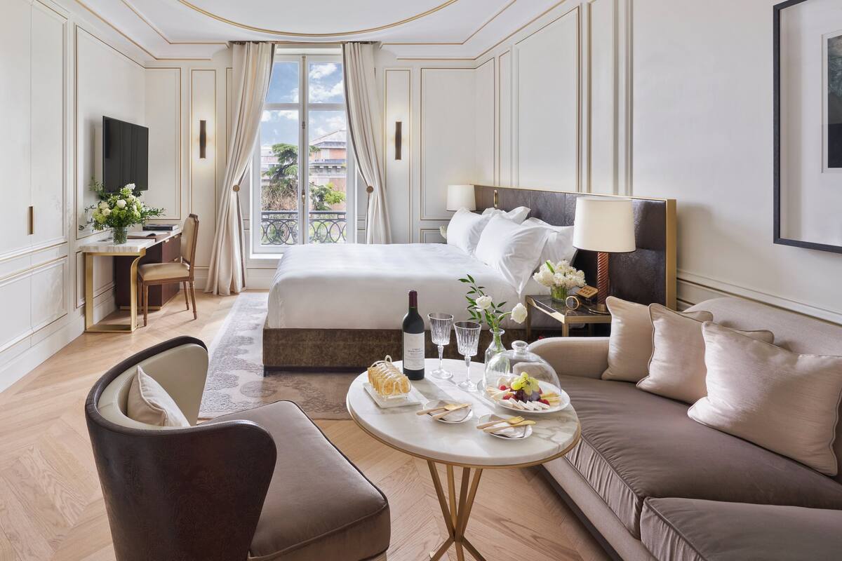 A hotel room with a large bed, a sofa, a desk with a computer, a TV, and a dining table with food and drinks.