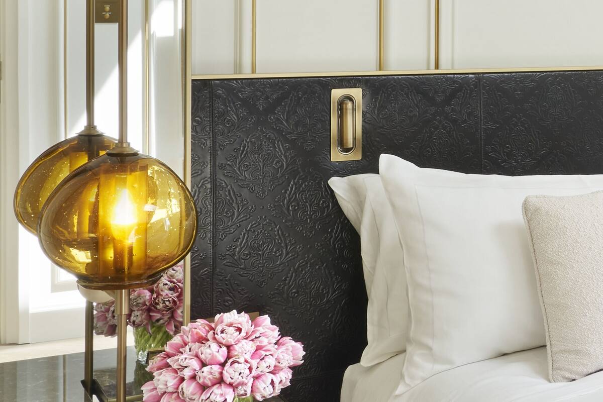 A bed with white linens and a single beige pillow, a black textured headboard, and a gold pendant light.