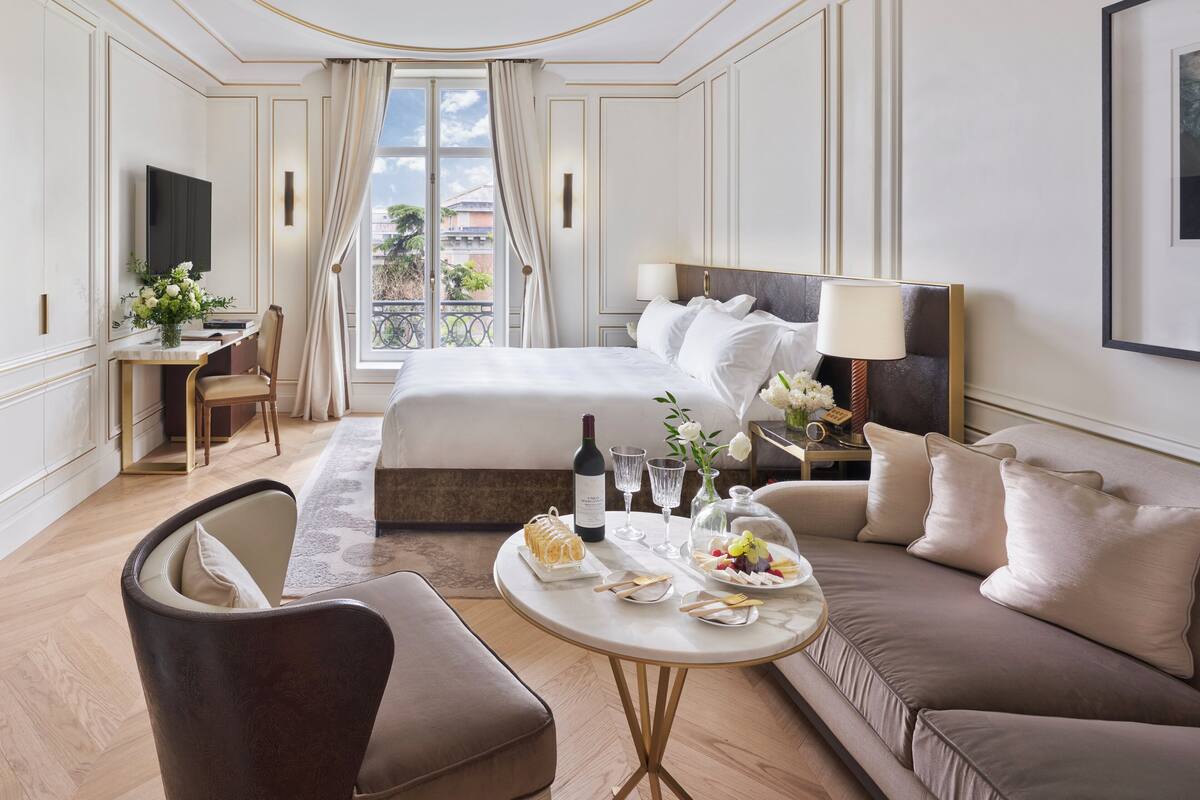 A hotel room with a large bed, a sofa, a desk with a computer, a TV, and a dining table with food and drinks.