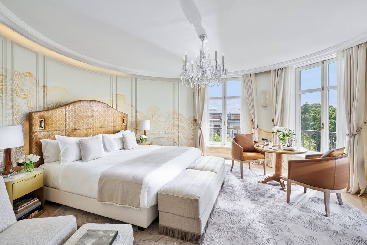 A spacious hotel room with a large bed, a sitting area with a table and chairs, and a view of the outdoors through large windows.
