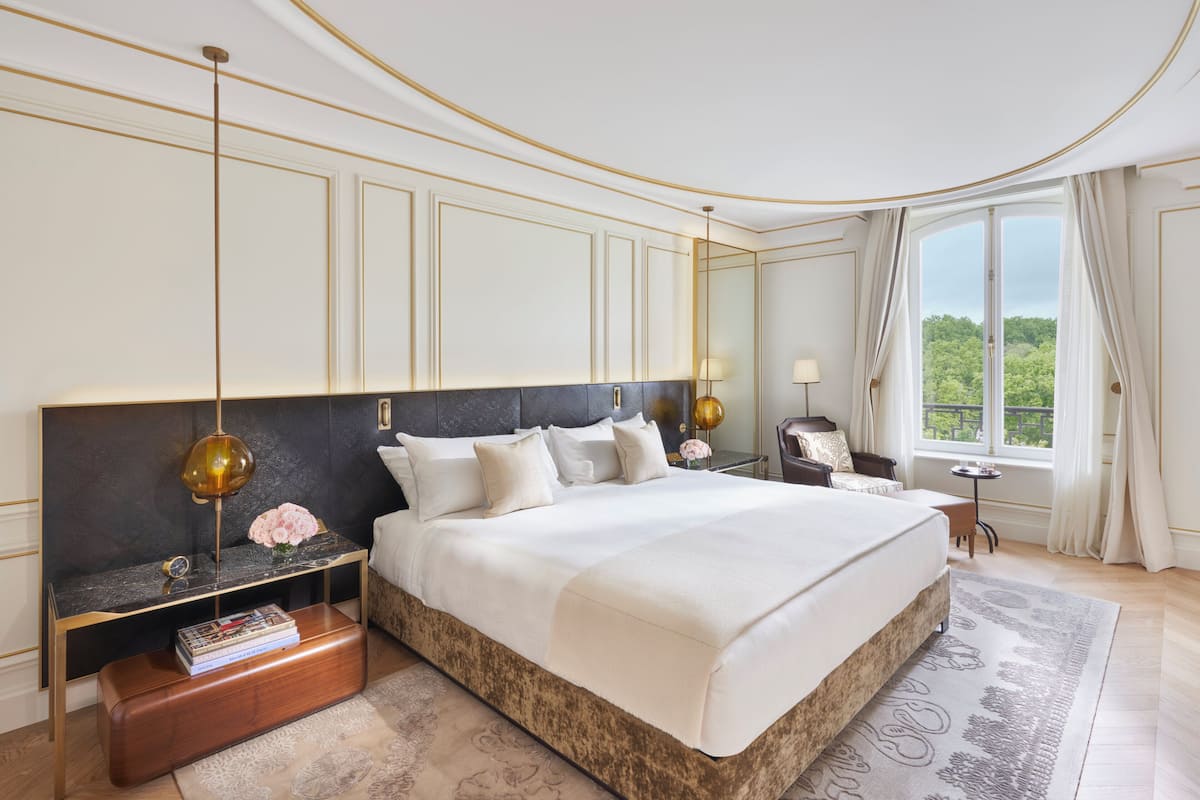 A spacious bedroom with a large bed, a nightstand with a lamp, a bedside table with a book, and a view of the outdoors through a large window.