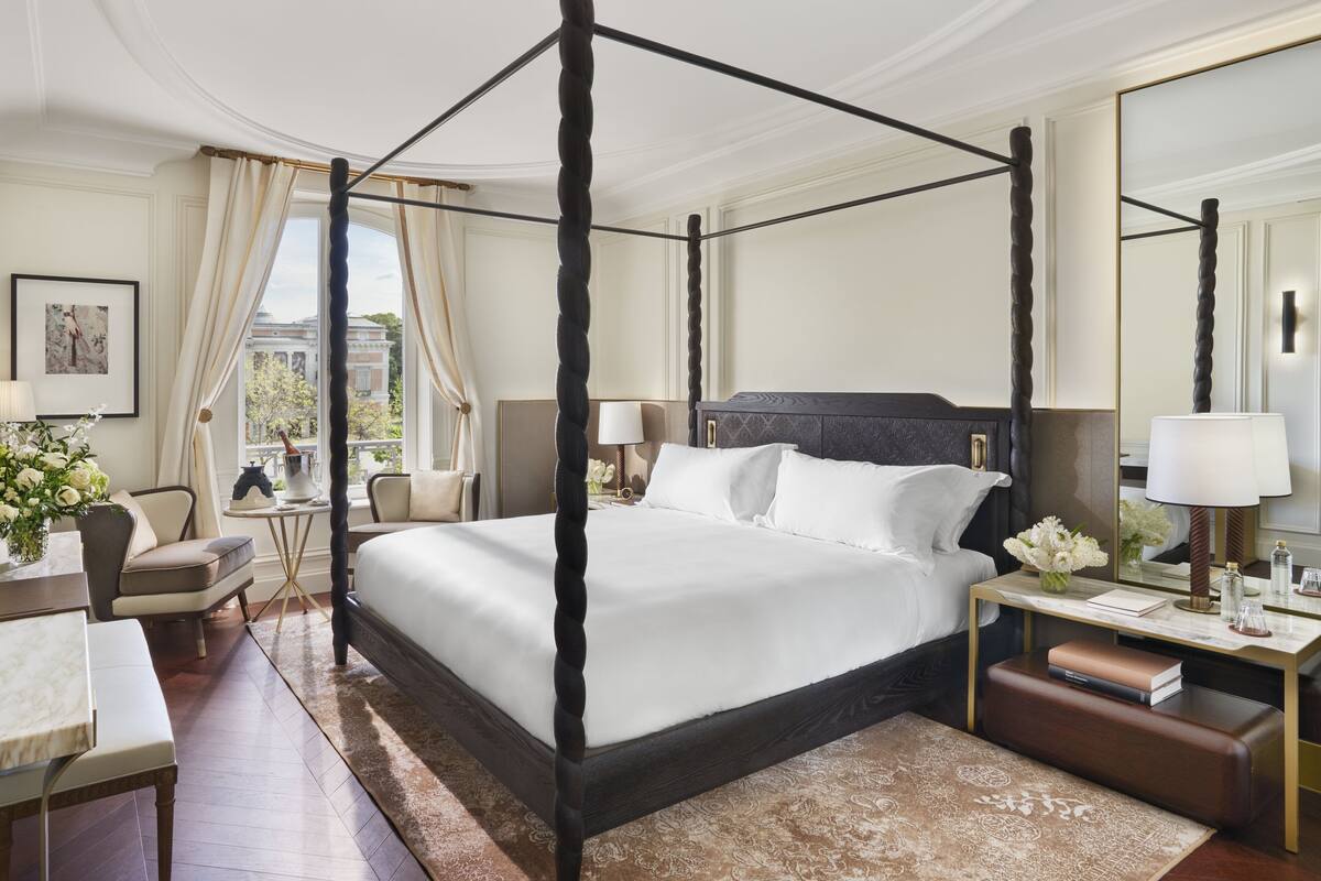 A four-poster bed with a dark headboard, a bedside table with a lamp, a chair, and a desk with a lamp.