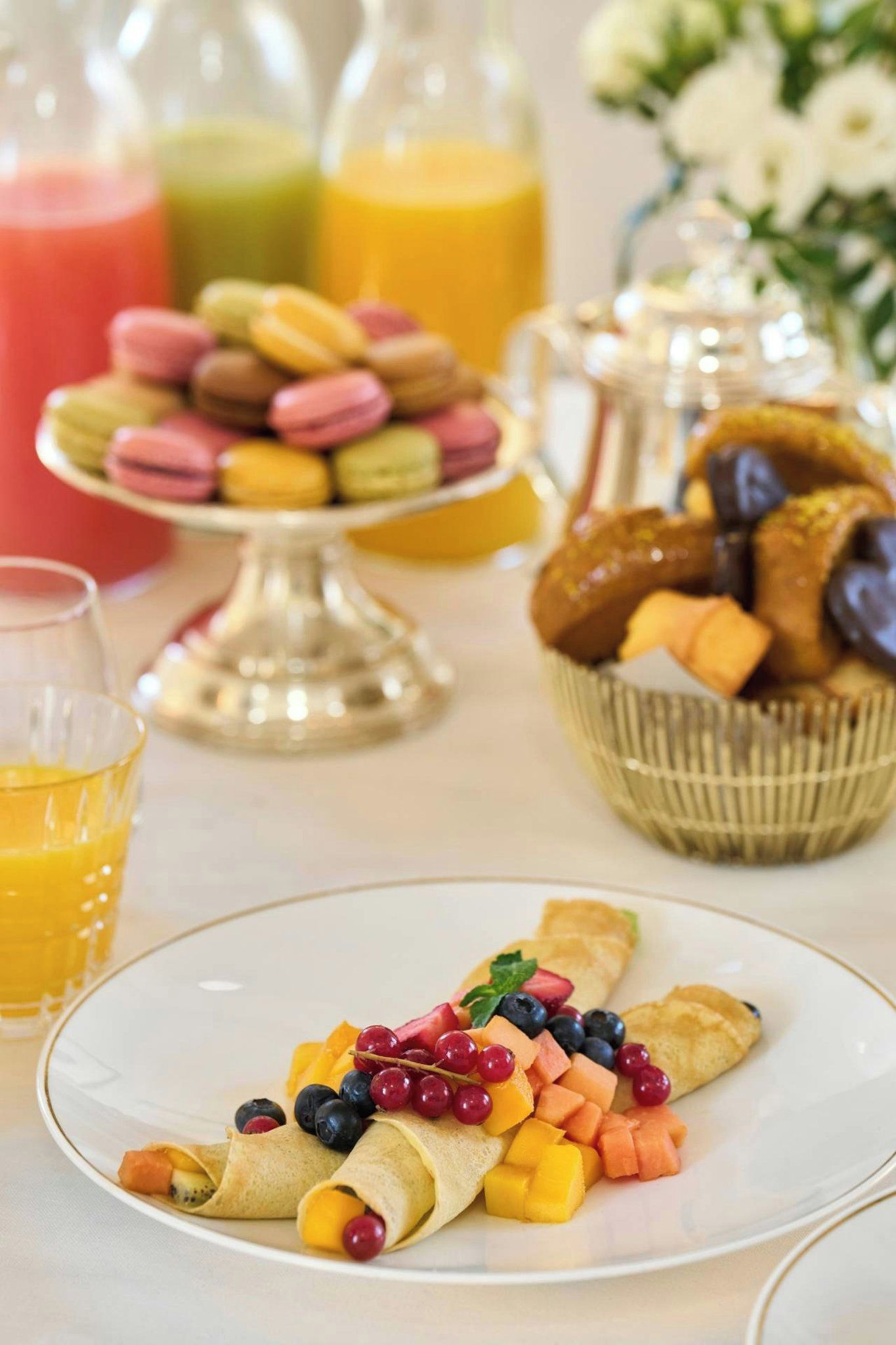 Un desayuno con crepes cubiertas de fruta, un vaso de jugo de naranja y una variedad de pasteles y galletas al fondo.