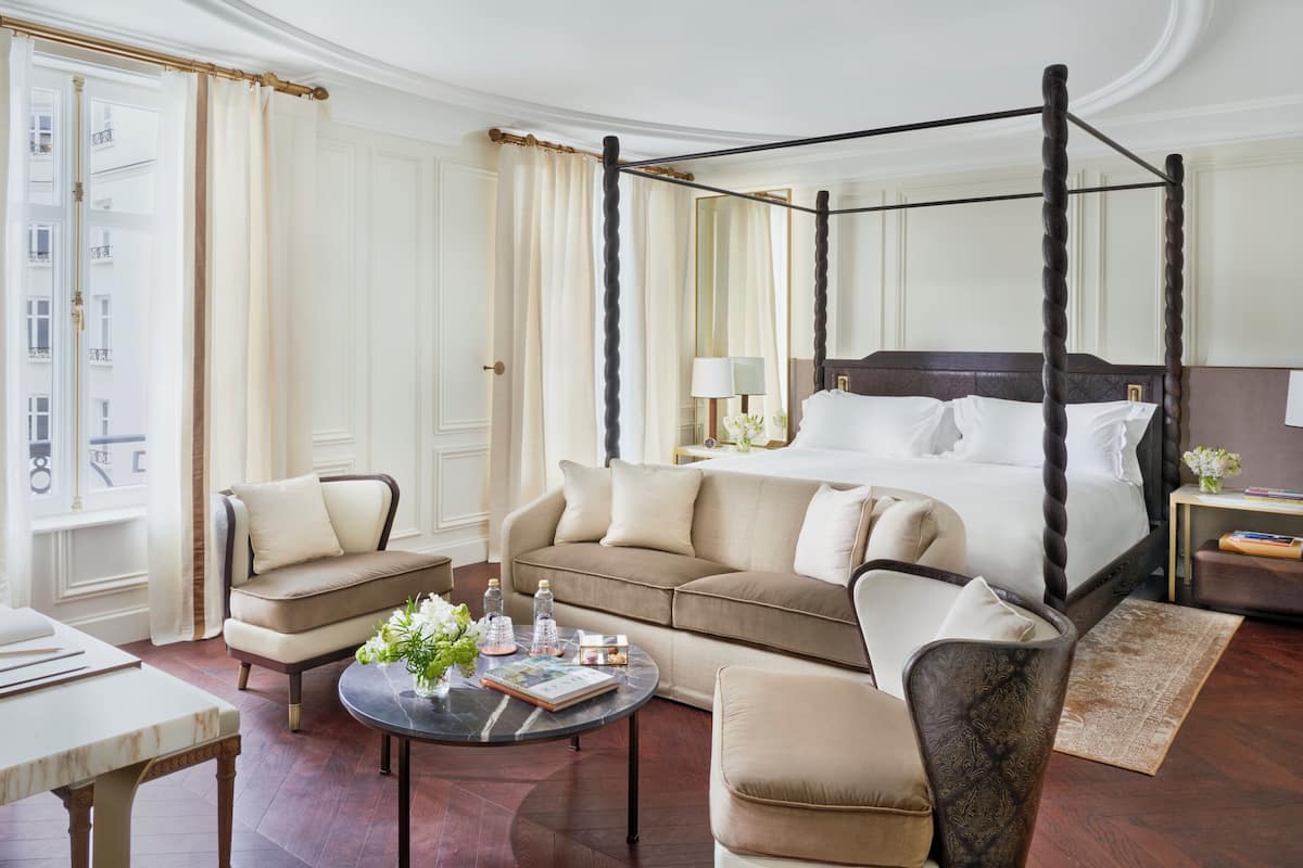 A four-poster bed with white linens, a beige sofa, a round coffee table with books and flowers, and a small side table with a lamp and a book.