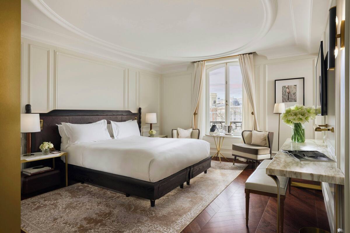 A hotel room with a large bed, a desk, a chair, a small table, and a view of the city.