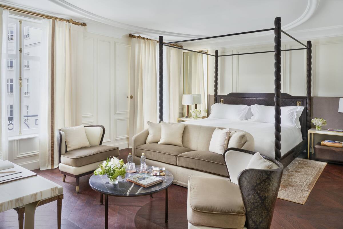 A four-poster bed with white linens, a beige sofa, a round coffee table with books and flowers, and a small side table with a lamp and a book.