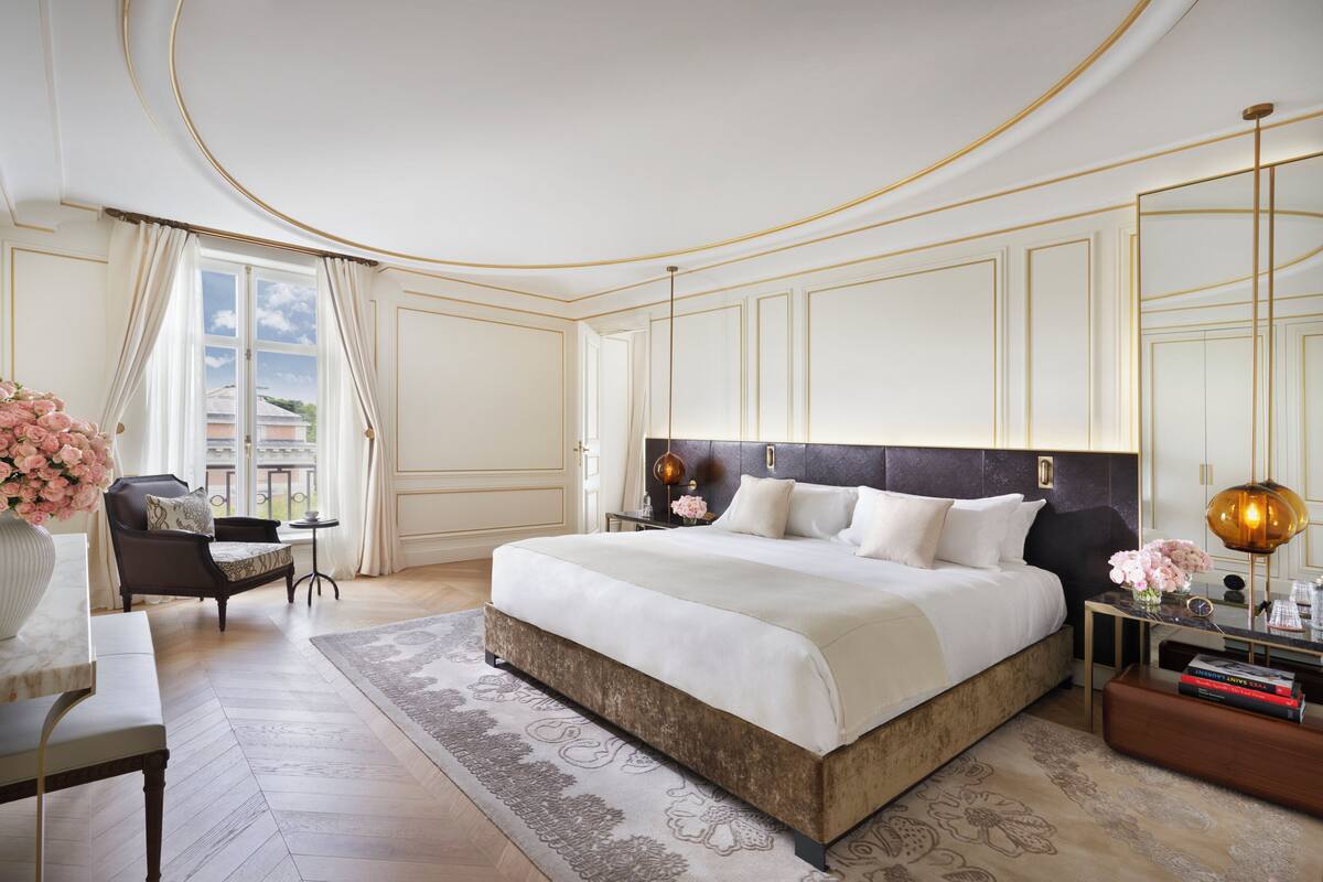 A spacious bedroom with a large bed, a sitting area with a chair and table, and a view of the outdoors through a window.