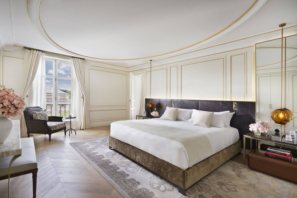 A spacious bedroom with a large bed, a sitting area with a chair and table, and a view of the outdoors through a window.