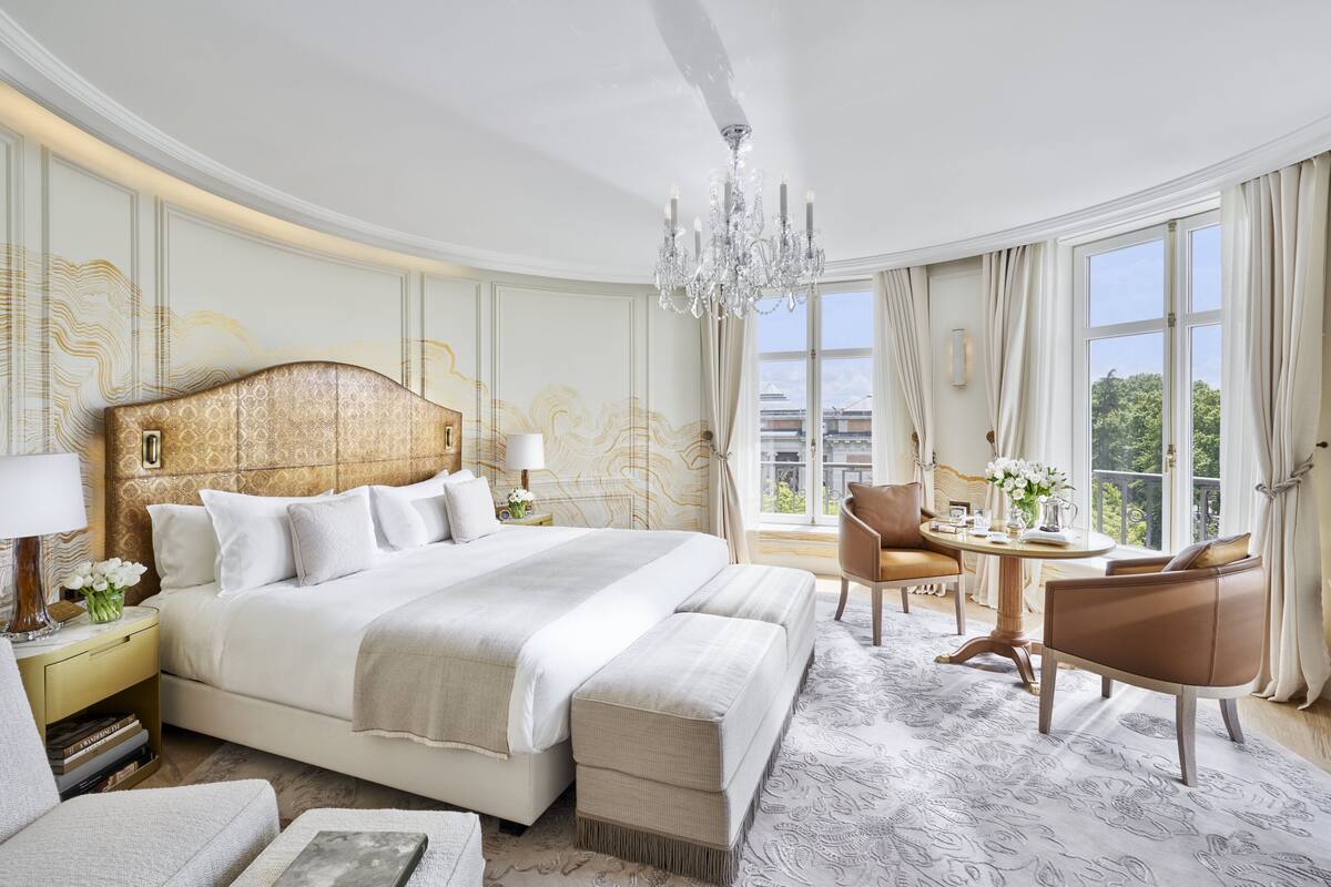 A spacious hotel room with a large bed, a sitting area with a table and chairs, and a view of the outdoors through large windows.