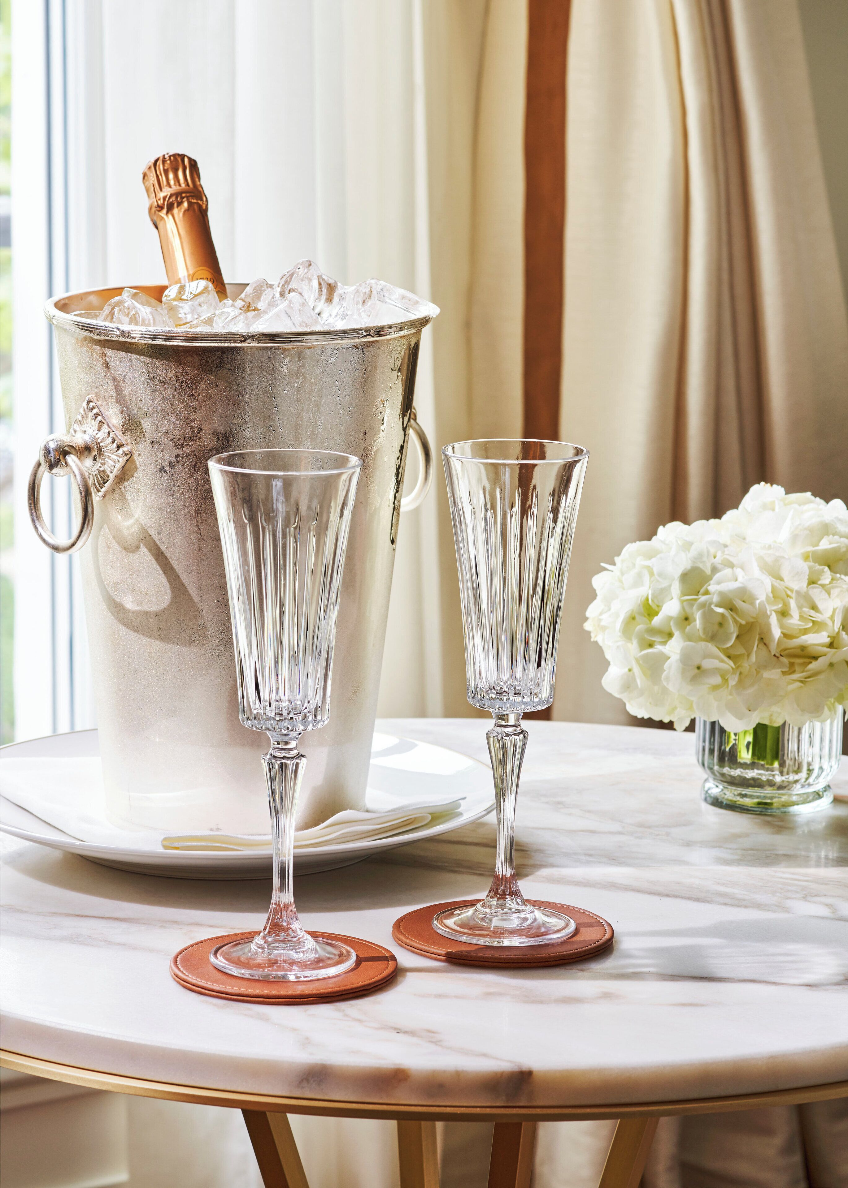 Un balde plateado para hielo con una botella de champán, dos copas de tallo y un jarrón con flores blancas sobre una mesa de mármol.