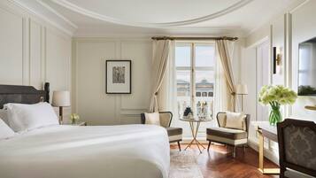 A hotel room with a large bed, two armchairs, a small table, and a view of buildings through a window.