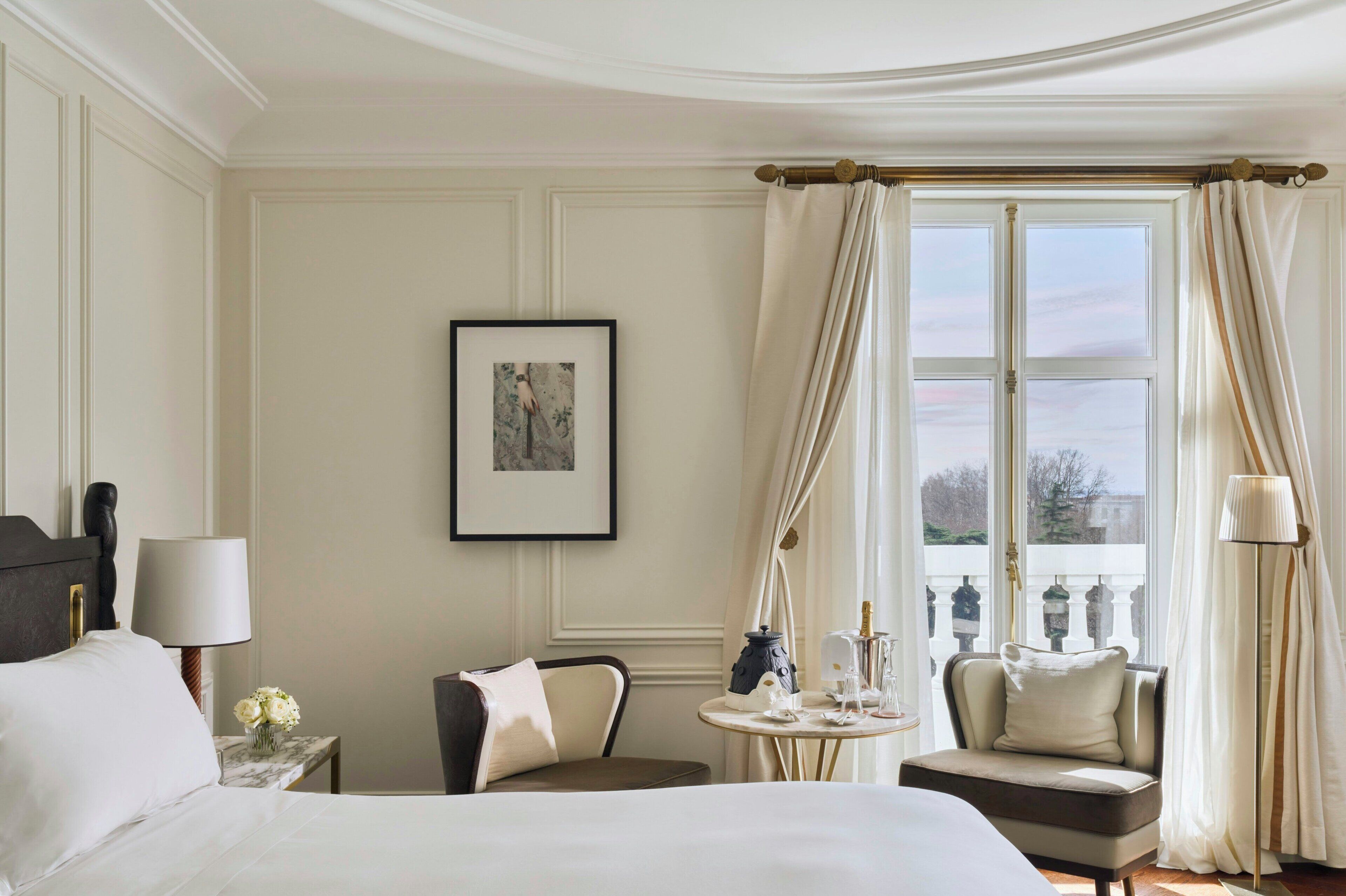 A hotel room with a bed, a chair, a small table with a lamp, a picture on the wall, and a window with curtains.