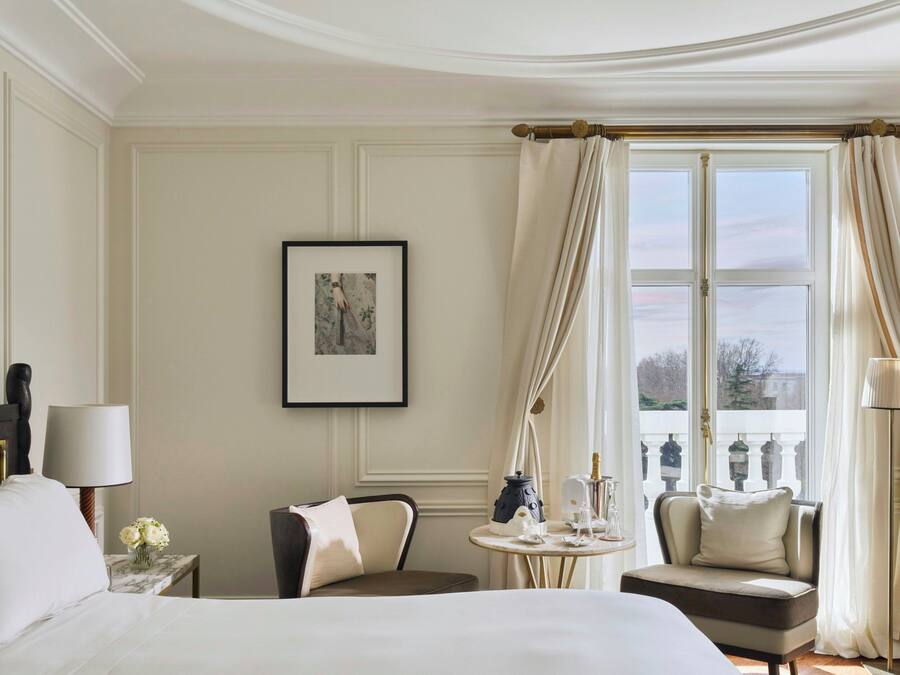 A hotel room with a bed, a chair, a small table with a lamp, a picture on the wall, and a window with curtains.