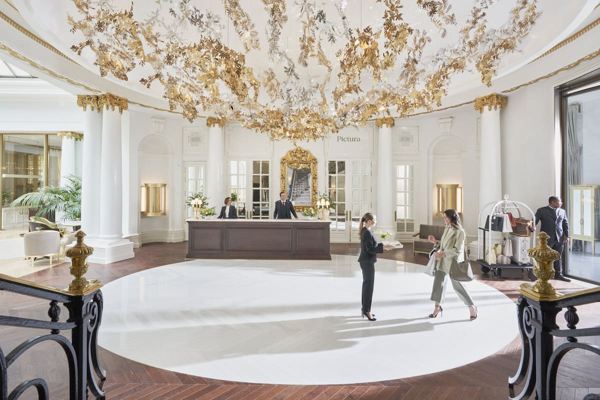 A grand lobby with a large white reception desk, ornate ceiling decorations, and classical architecture.