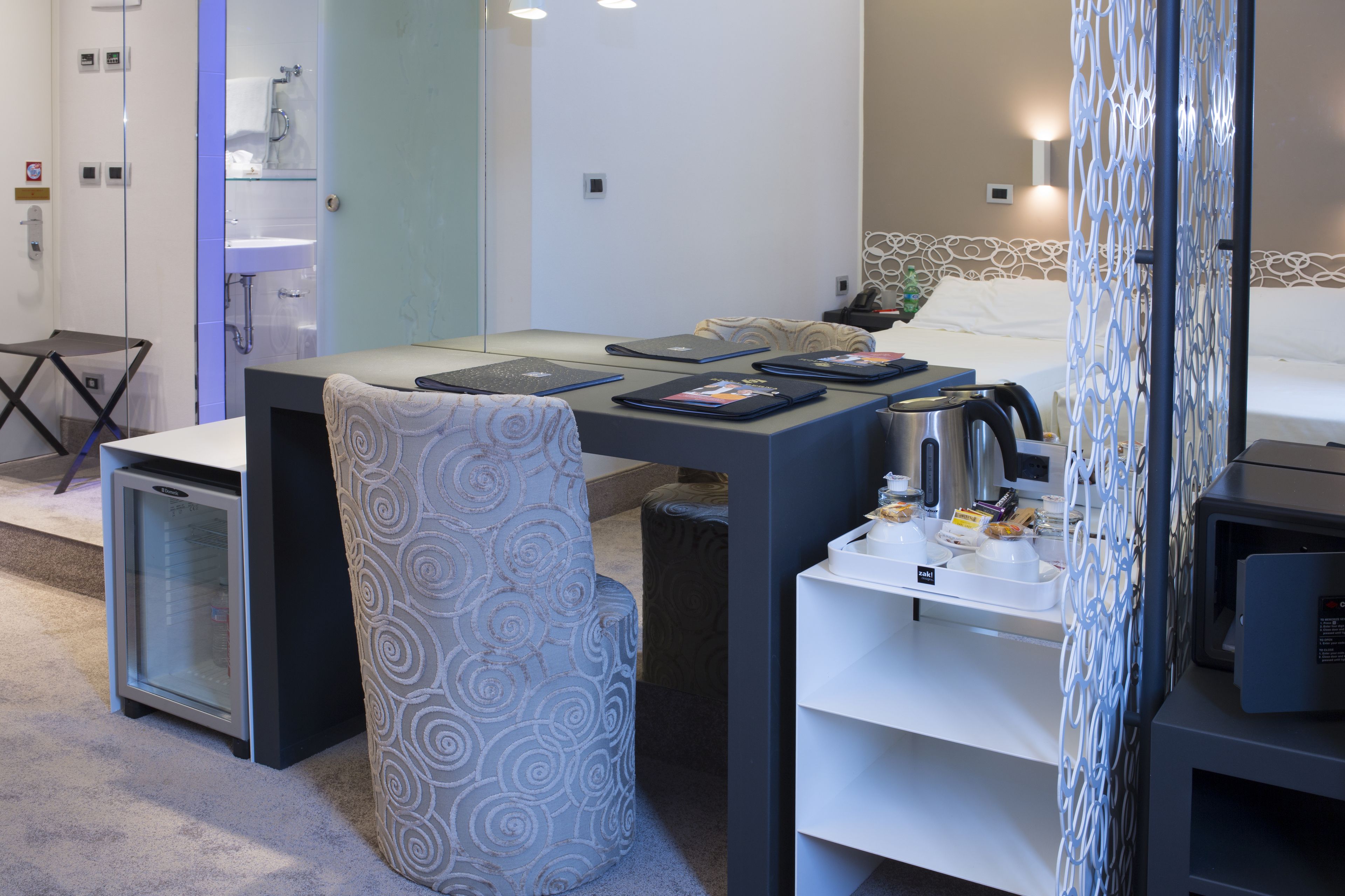 A compact hotel room with a bed, a desk with a laptop, a chair, a small refrigerator, and a microwave.