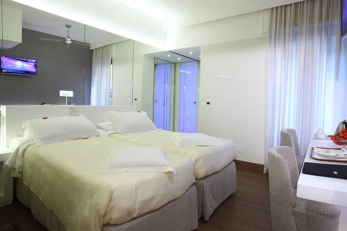 A modern hotel room with a large bed, a desk with a laptop, a television, and a mirror.