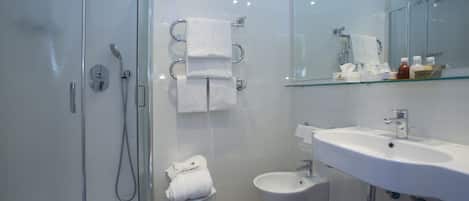 Un baño moderno con cabina de ducha de vidrio, un bidé, un lavabo blanco y una repisa de vidrio con artículos de aseo.
