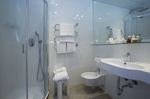 Un baño moderno con cabina de ducha de vidrio, un bidé, un lavabo blanco y una repisa de vidrio con artículos de aseo.