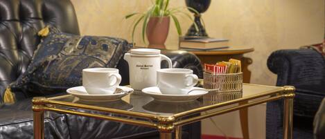 A living room with a leather sofa, a glass coffee table with a tray of white cups and saucers, a potted plant, and a lamp.