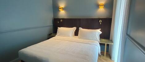 Una cama bien hecha en una habitación con paredes azules, dos mesitas de noche y luces fijadas a la pared.