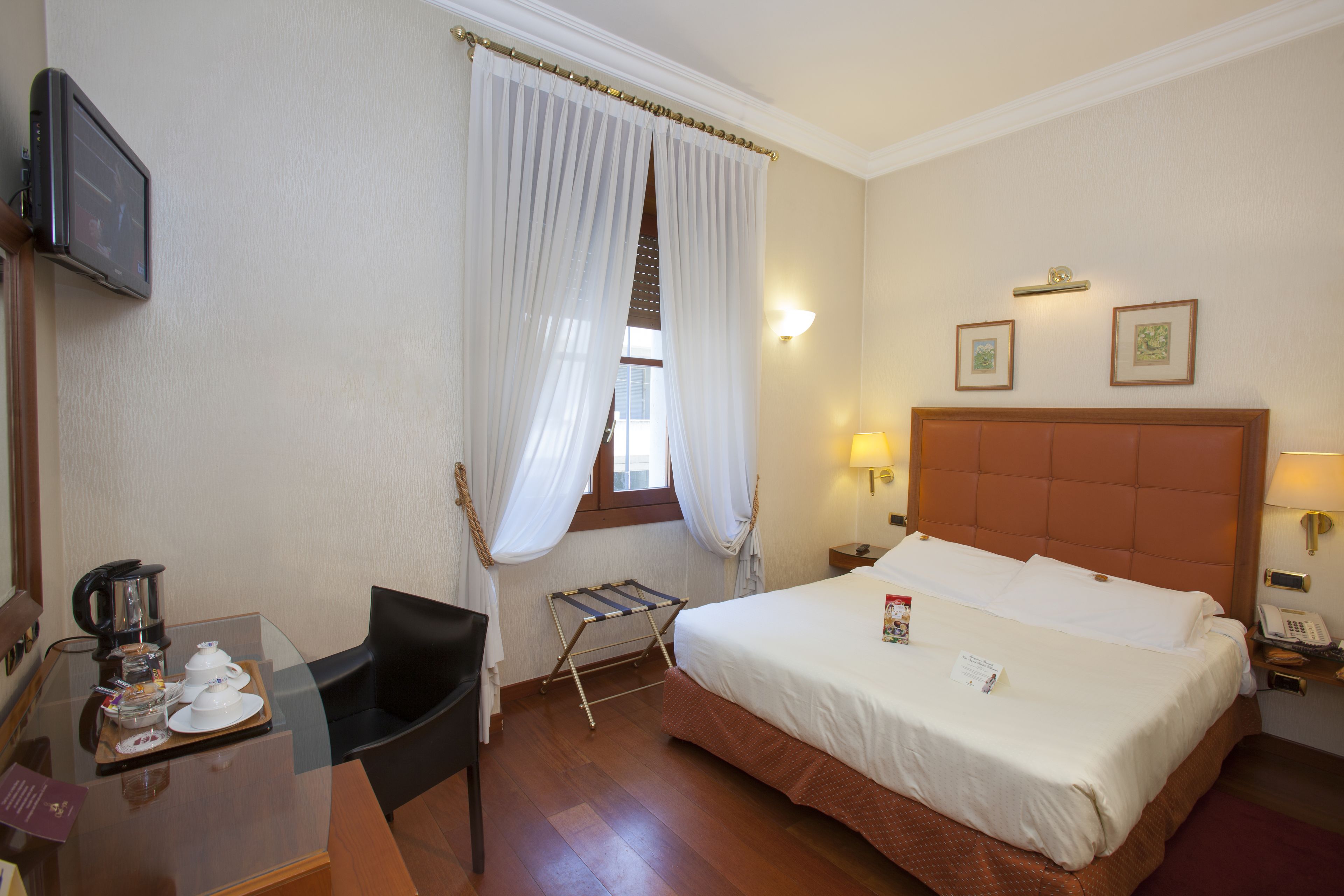 A hotel room with a bed, a desk with a coffee maker, a chair, a window with curtains, and a wall-mounted TV.
