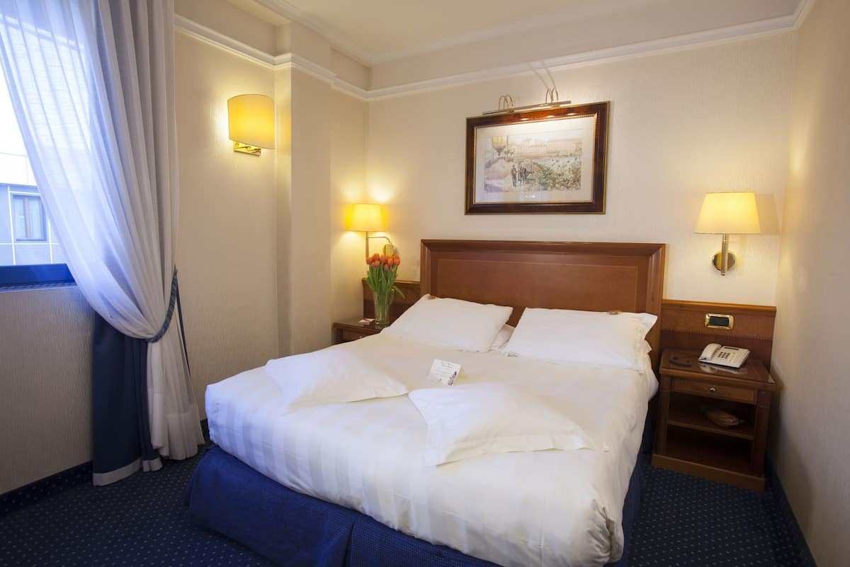A hotel room with a large bed, two bedside tables, a nightstand, a framed picture on the wall, and a window with curtains.