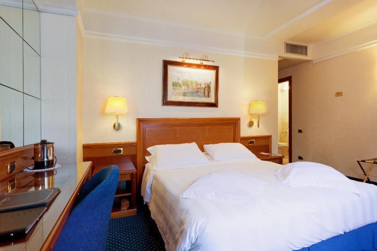 Habitación de hotel con cama, lámparas de noche, escritorio con silla y un cuadro en la pared.