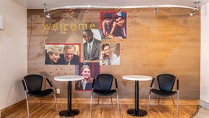 Un espace d'accueil avec une pancarte indiquant « Bienvenue », un mur orné de photos et trois chaises noires disposées autour de deux tables blanches.