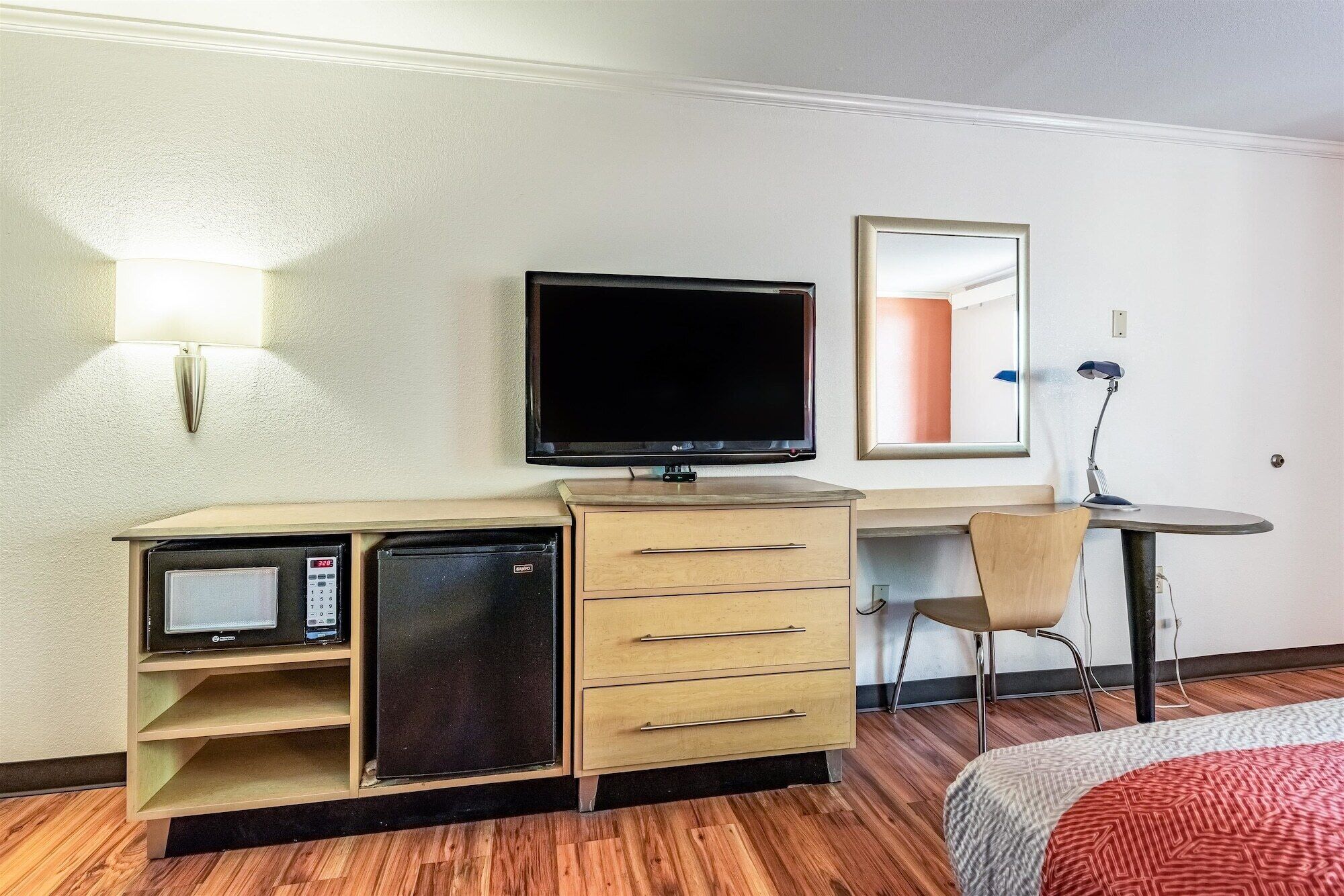 Une chambre d’hôtel compacte comprenant un bureau en bois, une télévision, un petit réfrigérateur, une chaise et un lit avec une couverture à motifs.