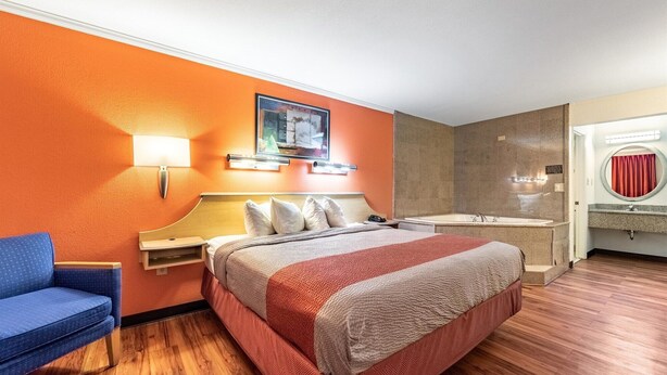 Une chambre d’hôtel avec un lit, une chaise bleue, un téléviseur fixé au mur, une salle de bain avec une baignoire et un rideau rouge.