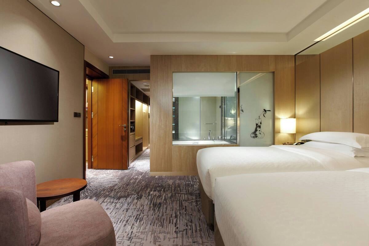 A hotel room with a large bed, a seating area, a TV, a bathroom with a glass shower, and a wooden wardrobe.