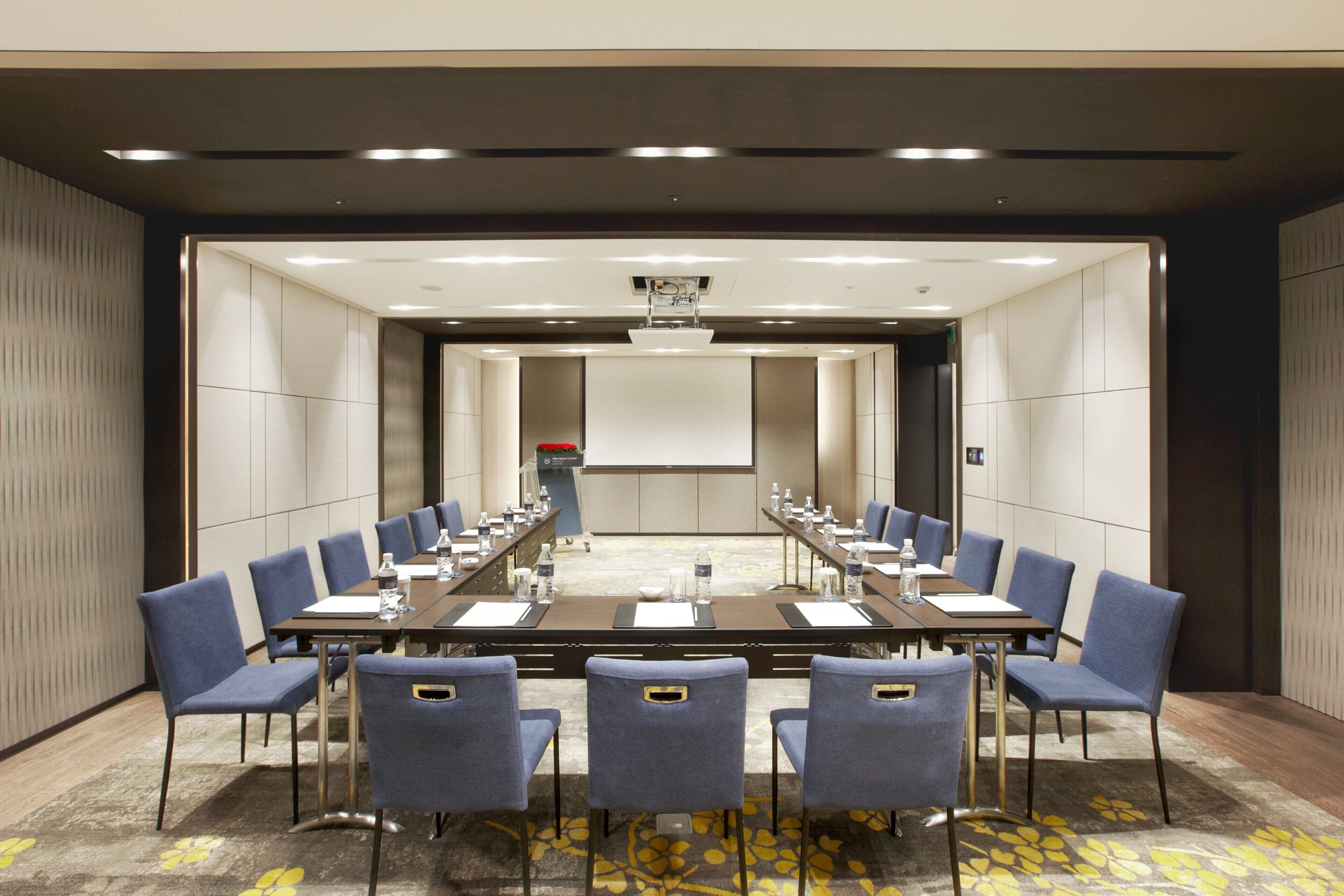 A conference room with a long table, blue chairs, and a projector screen at the front.