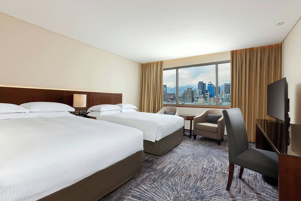 A hotel room with two beds, a desk, a chair, a television, and a large window with a city view.