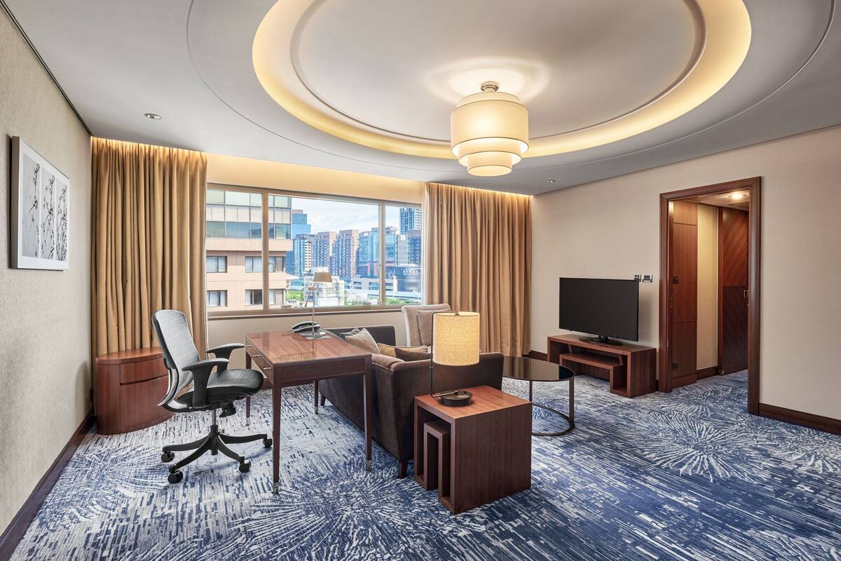 A hotel room with a large window, a desk with a chair, a sofa, a television, and a carpeted floor.