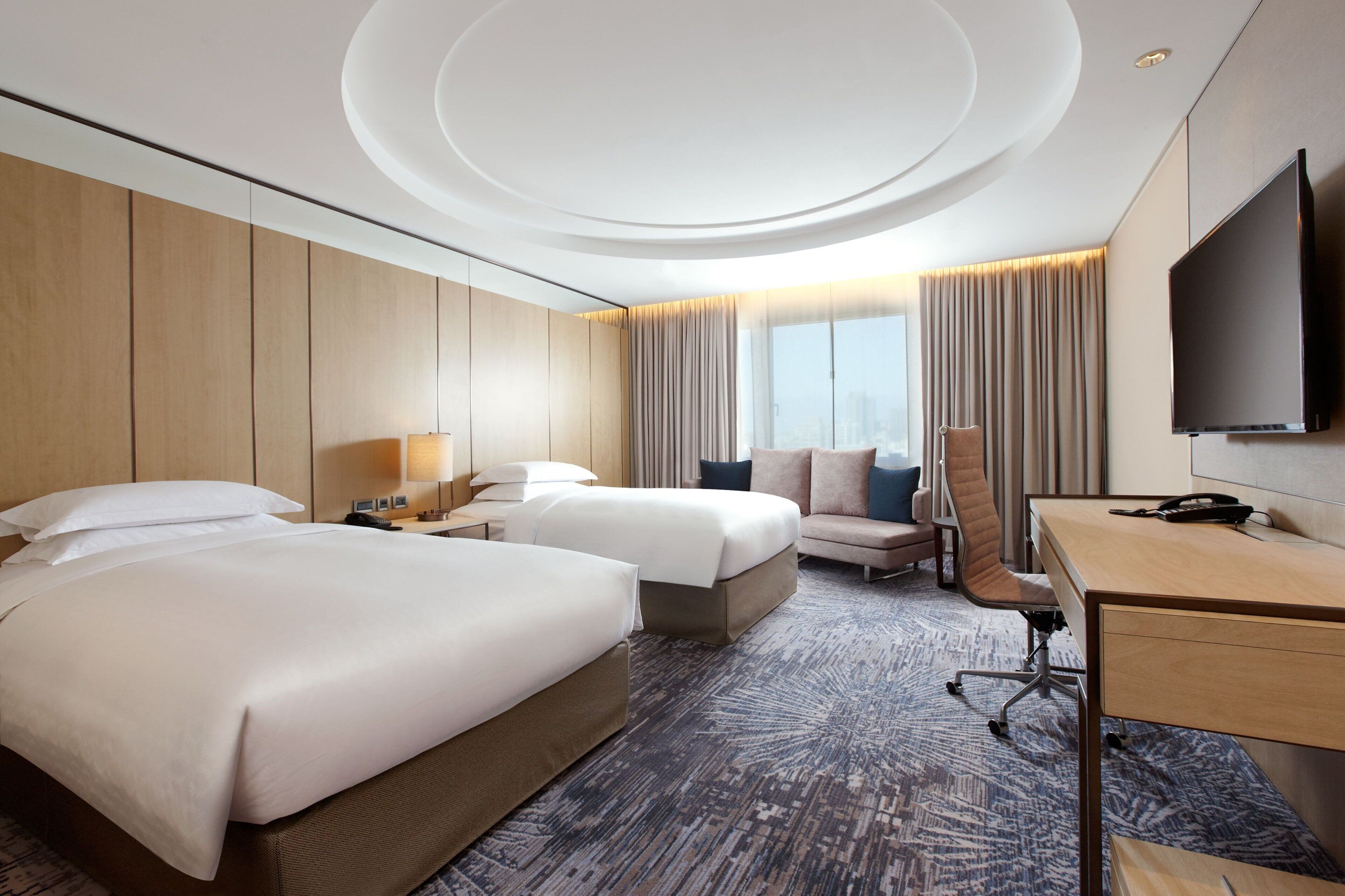 A hotel room with two beds, a desk, a chair, a TV, and a large window with curtains.