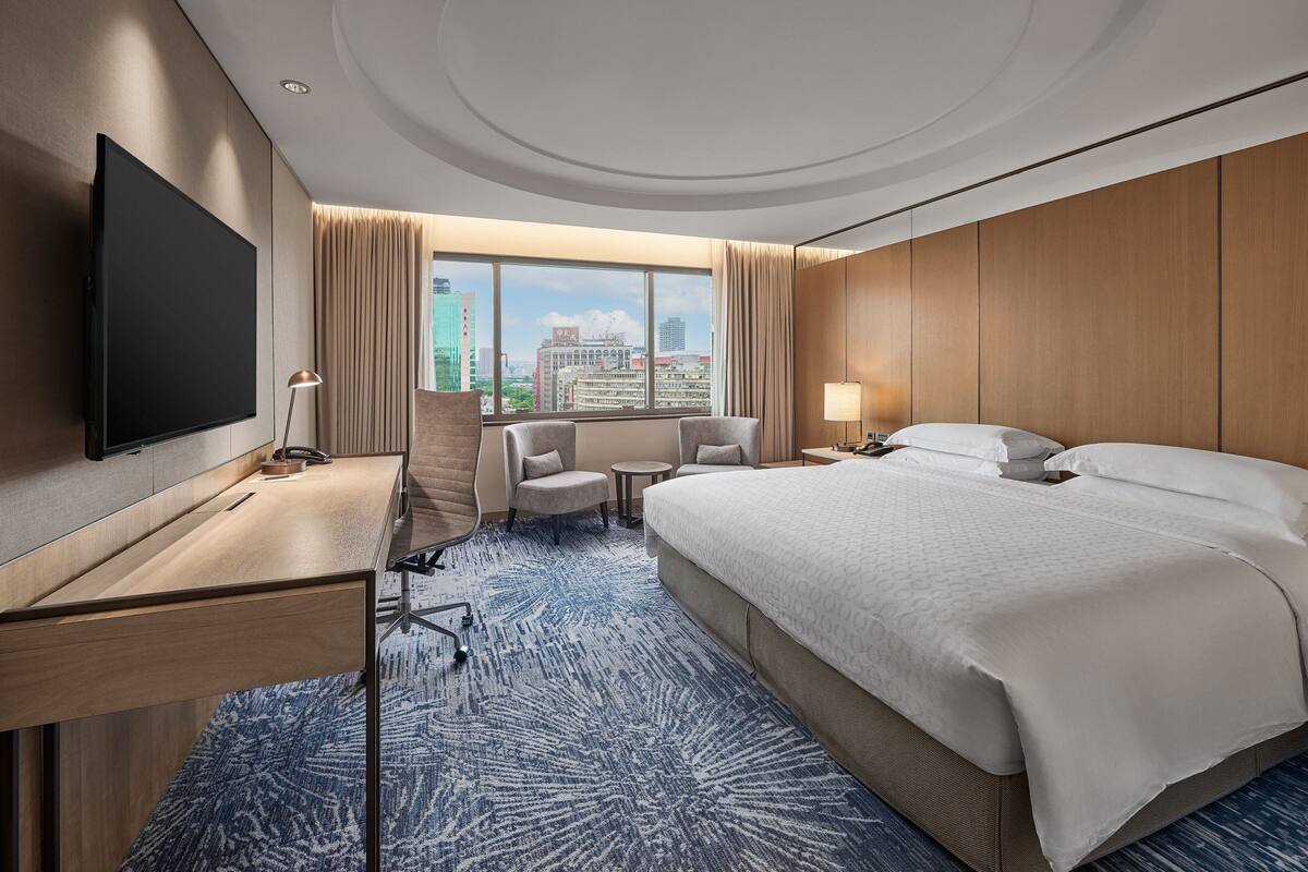 A hotel room with a large bed, a desk, a chair, a TV, and a view of the city.