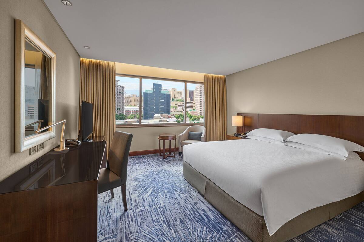 A hotel room with a large bed, a desk with a computer, a chair, a small table, and a window with a city view.