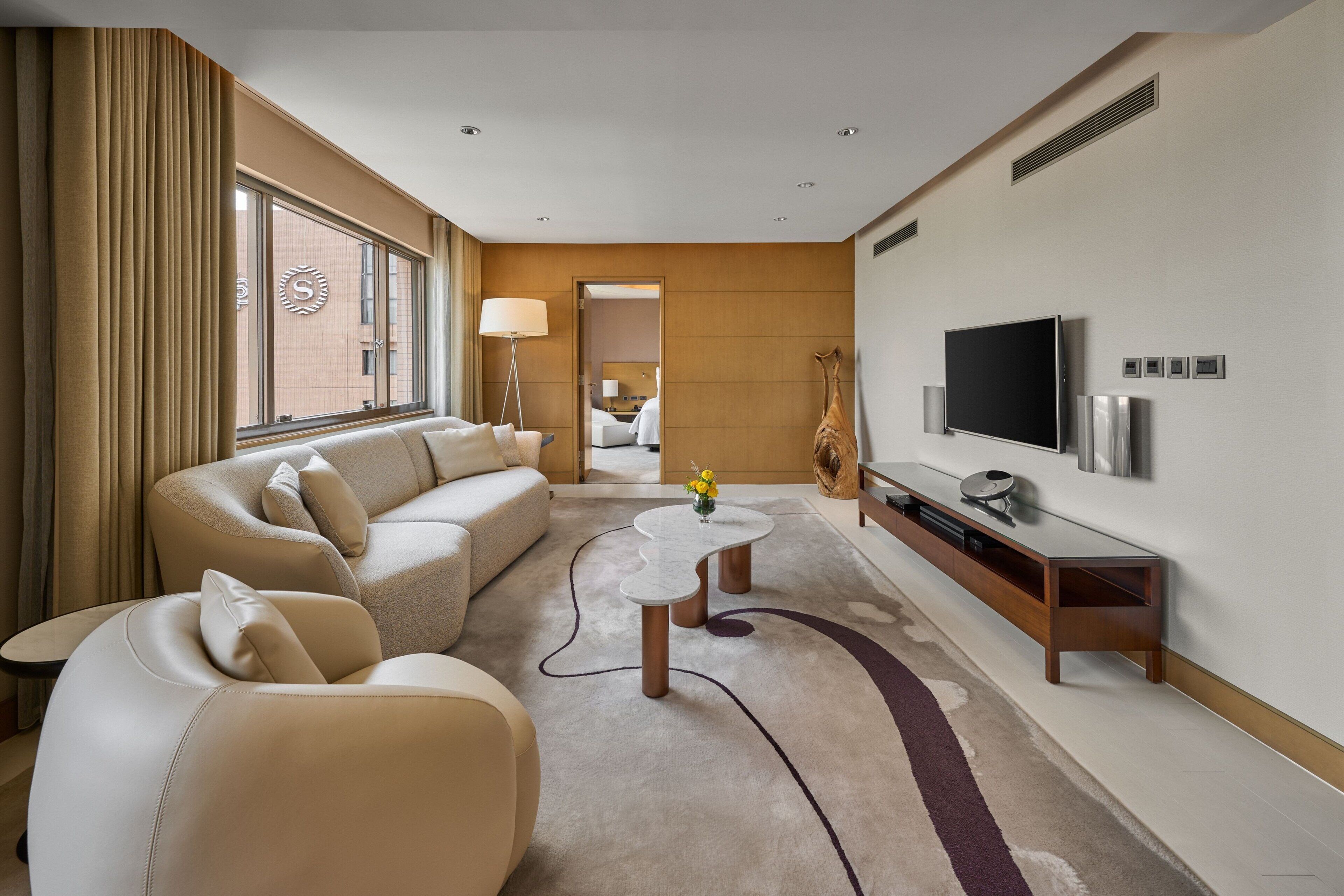 A modern hotel room with a sofa, a coffee table, a TV, and a large window.