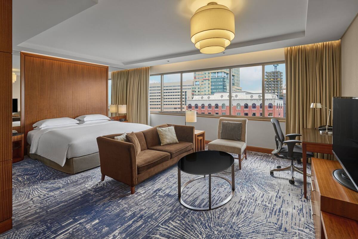 A hotel room with a large bed, a sofa, a desk, and a TV. There is a window with a view of buildings outside.