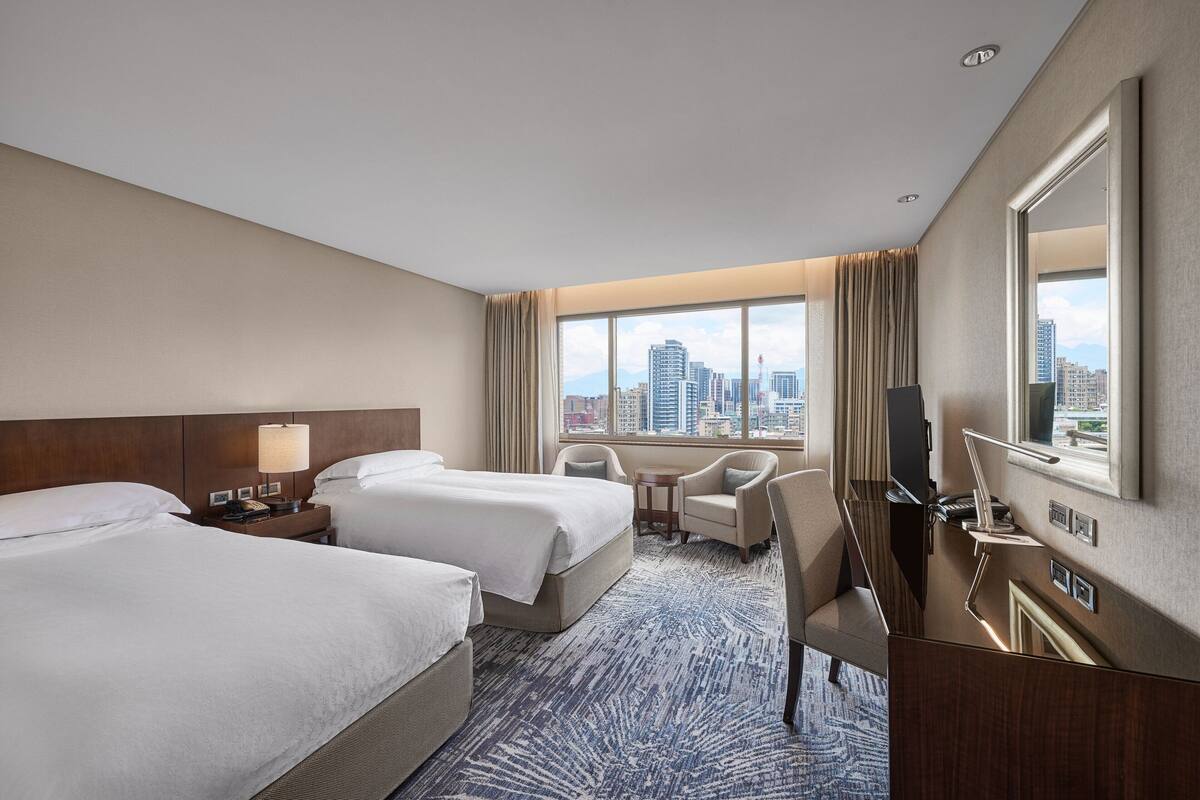 A hotel room with two beds, a desk, a chair, a TV, and a large window with a city view.