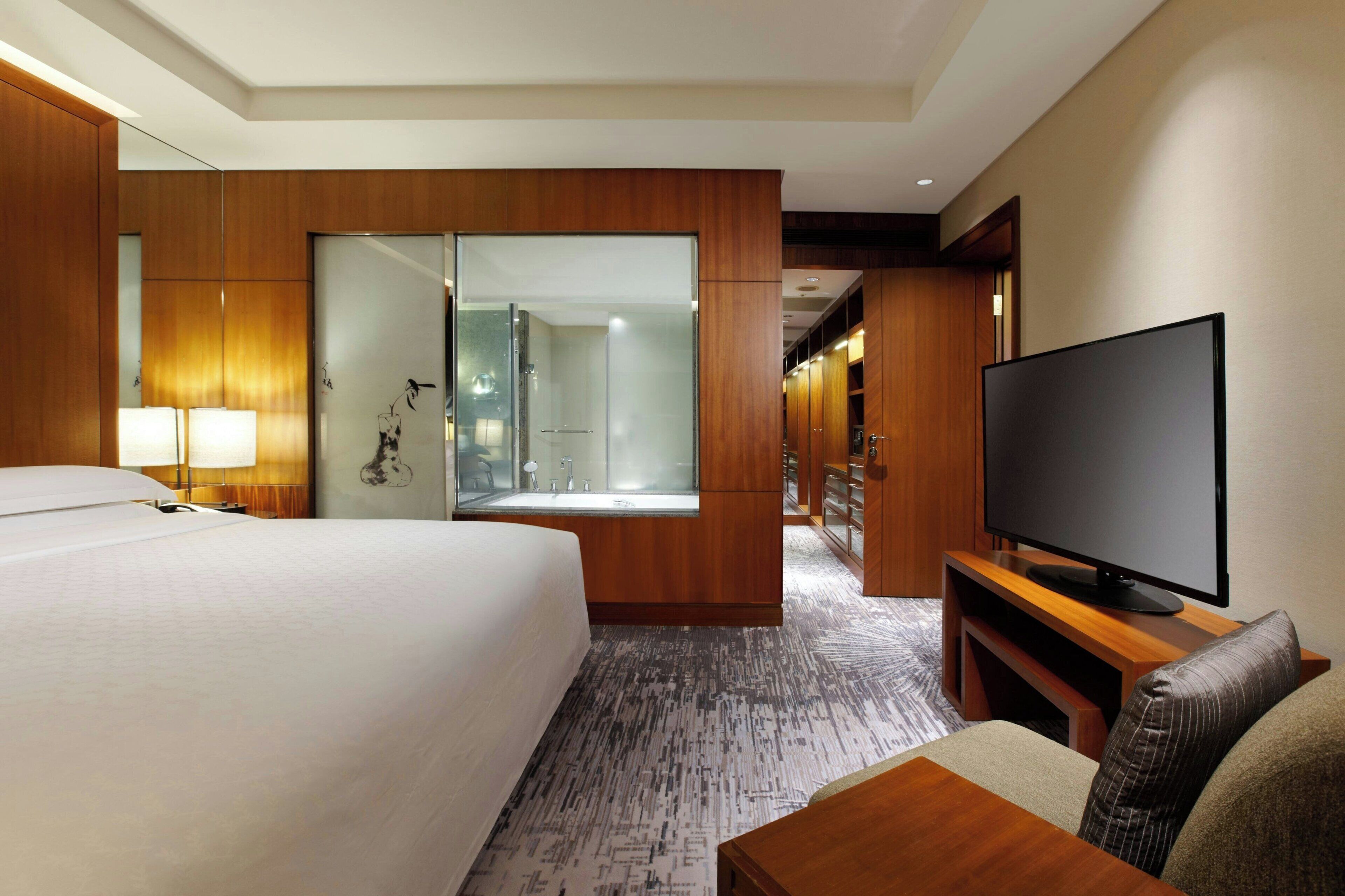 A hotel room with a large bed, a television, a mirror, and a bathroom with a glass shower.