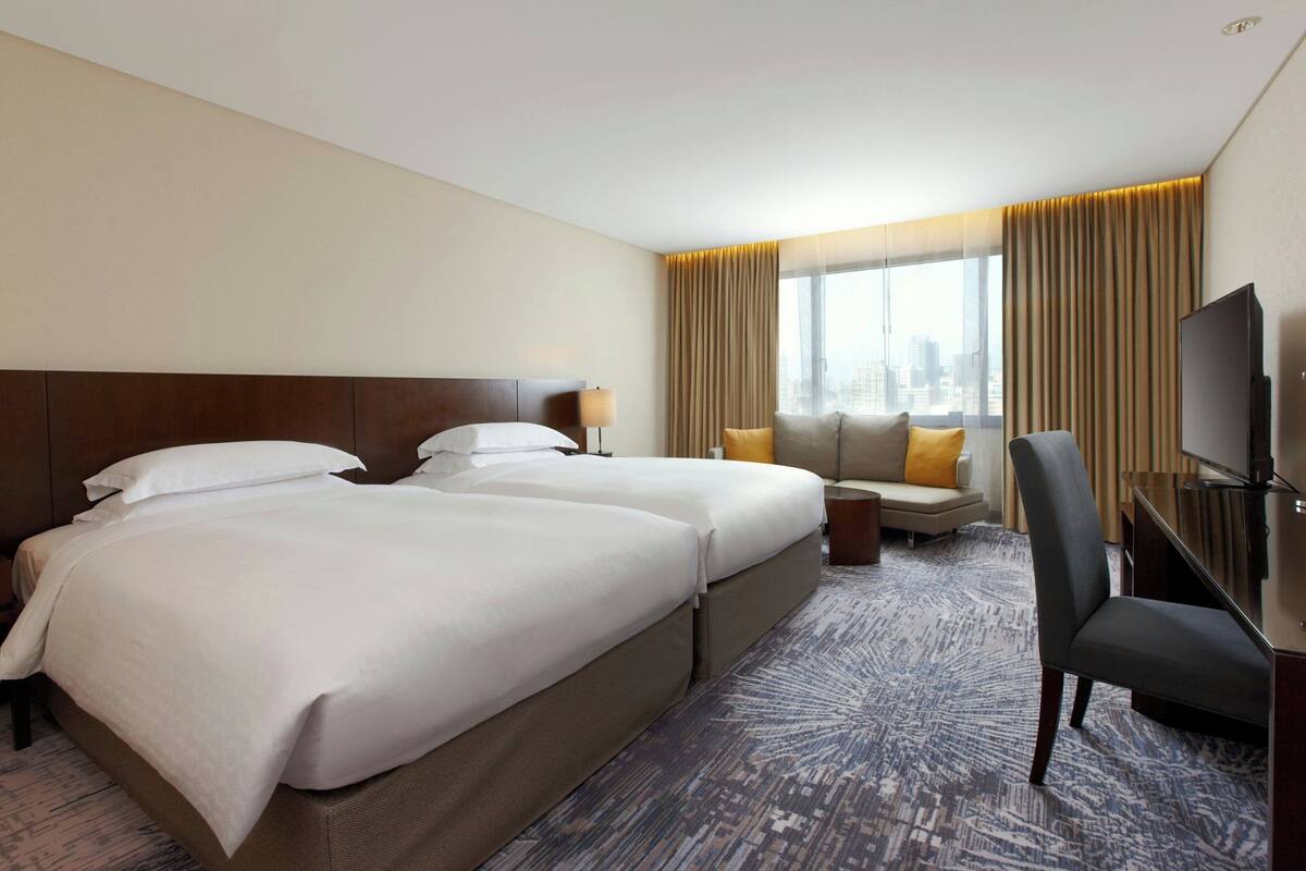 A hotel room with two beds, a sofa, a TV, and a city view.