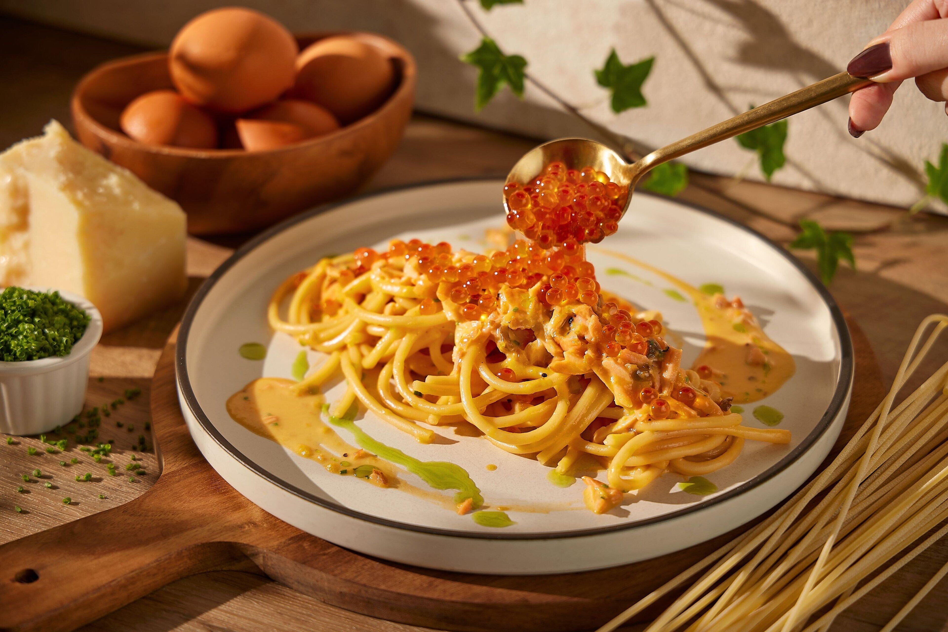 A plate of spaghetti with red caviar and sauce, a bowl of eggs, and a wooden cutting board with green herbs.