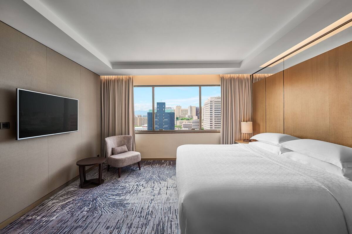 A hotel room with a large bed, a TV mounted on the wall, a chair, and a view of the cityscape through the window.