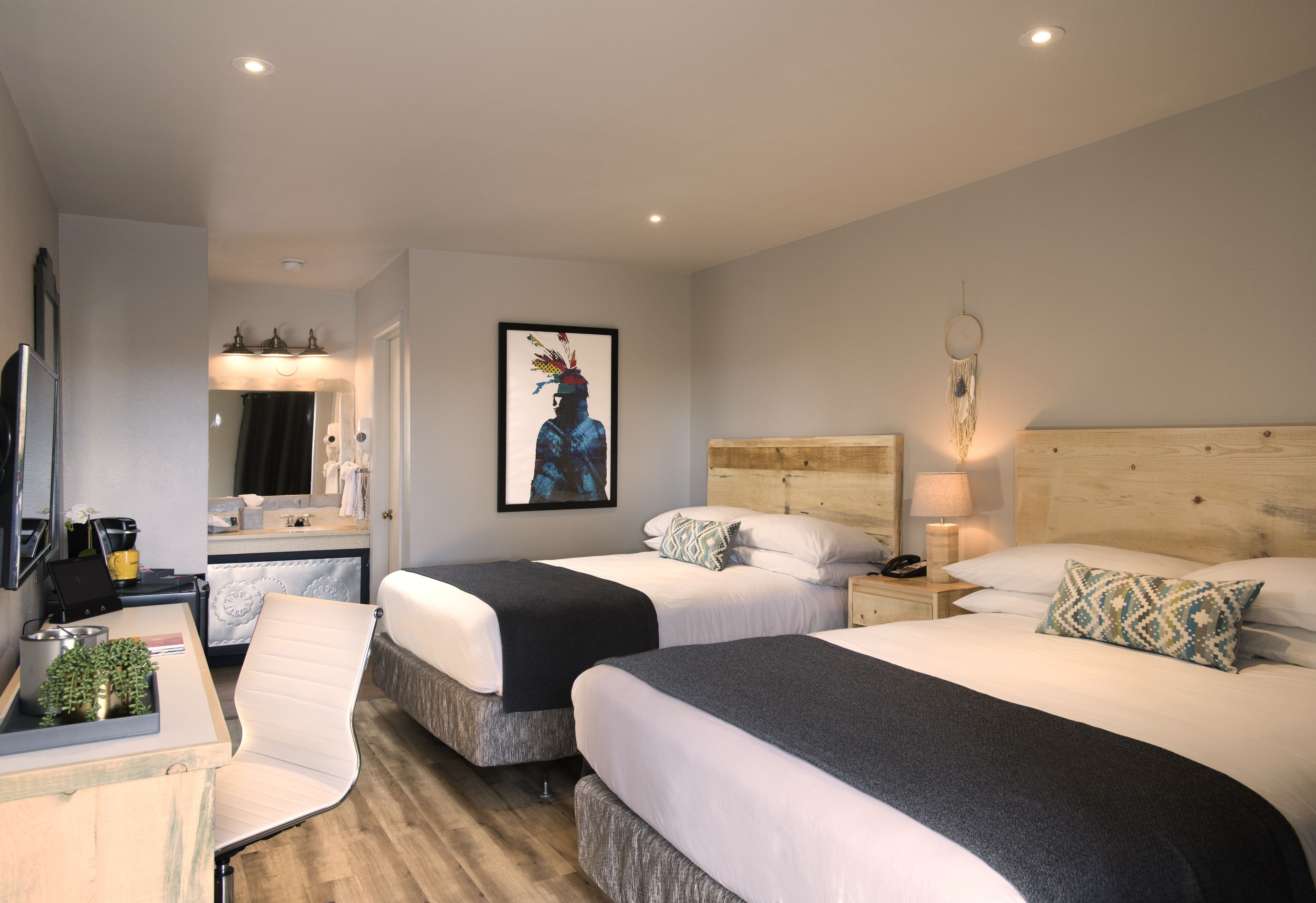 A hotel room with two beds, a desk with a computer, a chair, a small table with a plant, and a wall-mounted artwork.