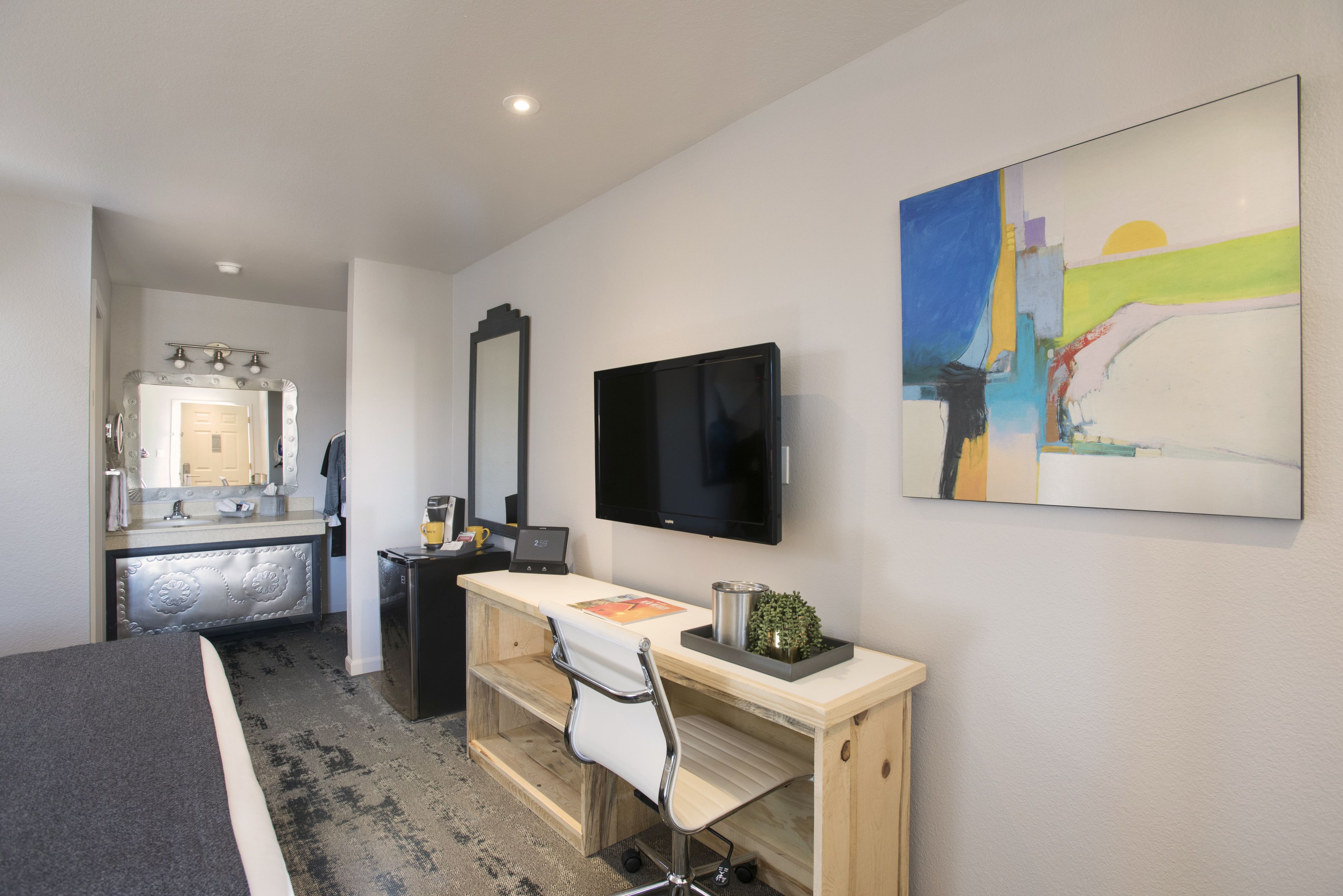 A hotel room with a bed, a desk with a chair, a television, a mirror, and a painting on the wall.