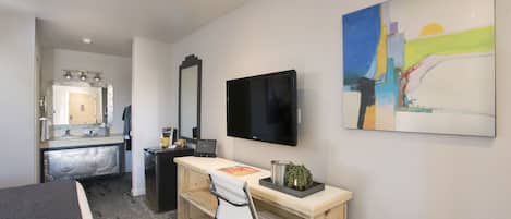 A hotel room with a bed, a desk with a chair, a television, a mirror, and a painting on the wall.