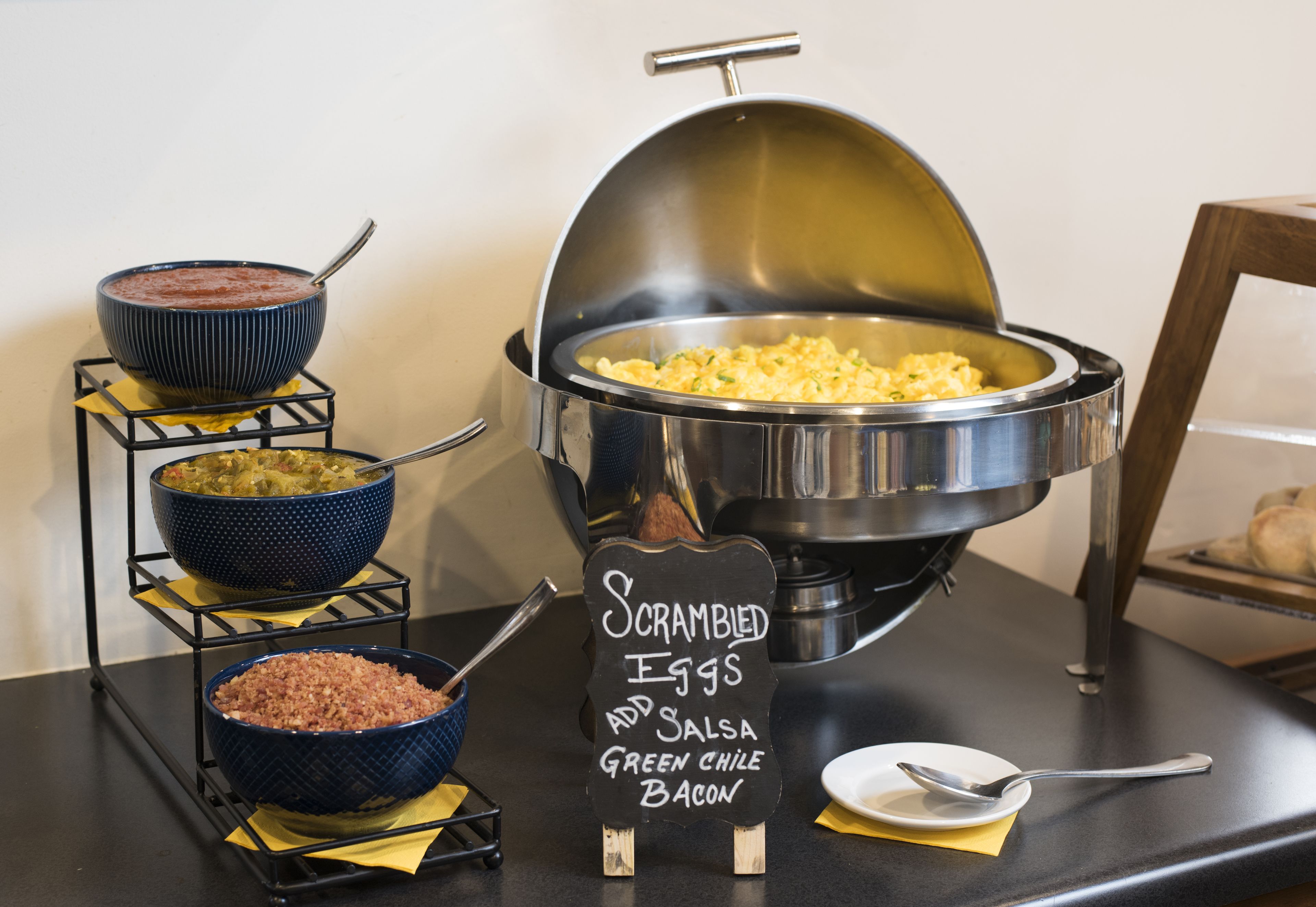 A buffet setup with scrambled eggs, salsa, green chile, and bacon.